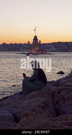 Muslimische Dame mit Telefon und Sonnenbrille sitzt auf Felsen und genießt den Sonnenuntergang über dem Bosporus Meer und dem Maidens Tower von Uskudar, Istanbul, Türkei Stockfoto