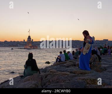 Die Menschen sitzen auf Felsen und genießen den Sonnenuntergang über dem Bosporusmeer und dem Maidens Tower von Uskudar, Istanbul, Türkei. Kreuzfahrtschiff hinten. Stockfoto