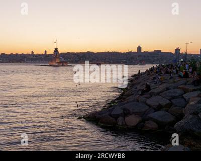 Die Menschen sitzen auf Felsen und genießen den Sonnenuntergang über dem Bosporusmeer und dem Maidens Tower von Uskudar, Istanbul, Türkei. Kreuzfahrtschiff hinten. Stockfoto