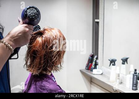 Friseur trocknet mit Haartrockner in einem Schönheitssalon, rotes kurzes Haar bei einem Teenager mit hellem Hintergrund. Alltag und Modetrends Stockfoto