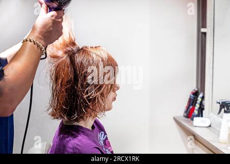 Friseur trocknet mit Haartrockner in einem Schönheitssalon, rotes kurzes Haar bei einem Teenager mit hellem Hintergrund. Alltag und Modetrends Stockfoto