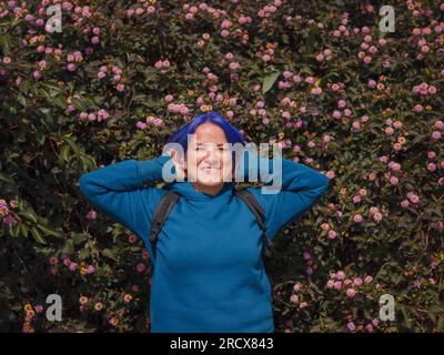 Eine Frau trägt einen hellblauen Hoodie und Jeans in Übergröße über der Blumenwand. Blauhaarige Frau draußen im Park in der Stadt Antalya. Kleidungsmodell, leerer Platz für Text o Design. Stockfoto