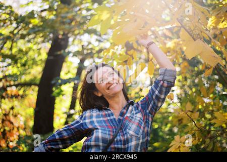 Lächelnde braune Frau mittleren Alters, die gelbe Herbstblätter bewundert Stockfoto