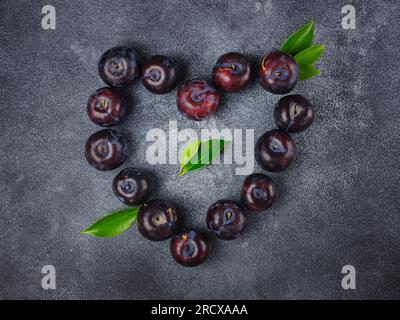 Süße Pflaumen auf dunklem Hintergrund. Frische blaue Pflaumen mit Blättern. Kulinarische Frucht im Hintergrund. Sommerpostkarte. Die Pflaumen werden im Garten geerntet und liegen in Herzform, Draufsicht Stockfoto