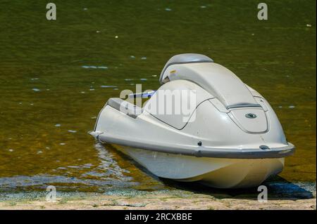 Ein weißer, einfacher und unmarkierter Jetski mit grauen Akzenten sitzt auf einer algenüberdachten Bootsrampe leicht im Wasser eines grünen, braunen Flusses oder Sees. Stockfoto