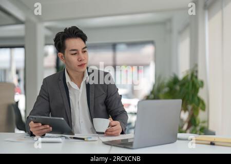 Fokussierter junger Geschäftsmann aus Asien, der am Schreibtisch sitzt und sicherstellt, dass sein Unternehmen genug Geld für die Finanzplanung verdient Stockfoto