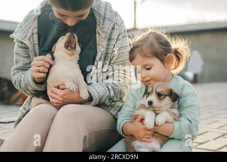 Eine schöne und glückliche junge Mutter mit ihrer kleinen Tochter, die Welpen im Hof streichelt. Komfort. Warme Töne. Hochwertiges Foto Stockfoto