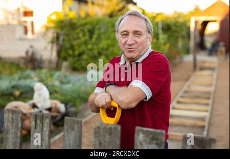 Ein erwachsener Mann stand entspannt neben dem Zaun, mit einer Schaufel im Garten, die lächelte Stockfoto