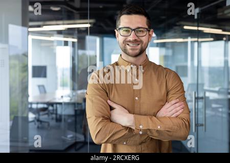 Porträt eines jungen männlichen Programmierers, kreativen Designers, Entwickler-Ingenieurs. Er steht im Büro, mit gekreuzten Armen auf der Brust und sieht mit einem Lächeln in die Kamera. Stockfoto