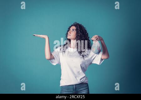 Studioaufnahme einer schönen, selbstbewussten, weißen, geschwungenen Modelin trägt blaue Jeans und ein legeres, einfarbiges weißes T-Shirt, isoliert auf blauem Hintergrund. P Stockfoto