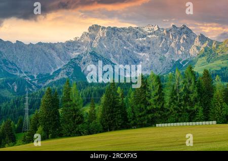 Malerischer Sonnenuntergang in den Alpen. Alpenpass zwischen Schweiz und Österreich. Grüne Alpenwiesen mit Heuballen und Stromleitungen mit hohen Bergen Stockfoto