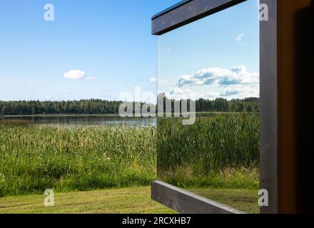 Detailansicht des modernen spiegelreflektierenden Fensters auf dem Gebäude inmitten der wilden Natur. Luxuriöses Cottage-Konzept. See, Schilf, Bäume, blauer Himmel. Stockfoto