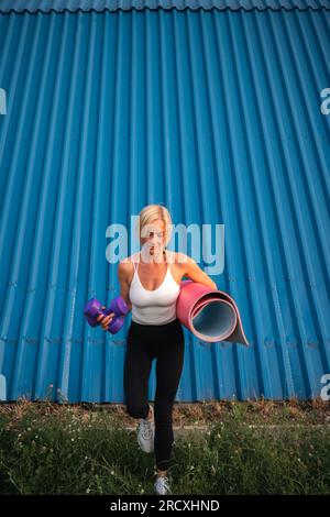 Blonde Frau mittleren Alters mit Trainingsmatte und Kurzhanteln, die am Sommertag bei Sonnenuntergang vor den Gebäuden spazieren Stockfoto
