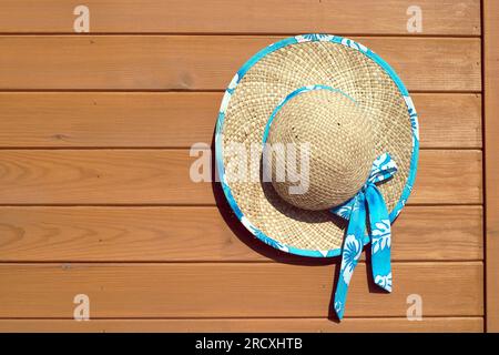 Sandig-brauner rustikaler weiblicher Strohhut mit dekorativen Blumen hängt an der Holzwand als Hintergrund Vorderansicht Stockfoto