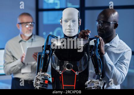 Afroamerikanischer Ingenieur, der Roboter mit Werkzeugen repariert, während sein Kollege ihn im Hintergrund programmiert Stockfoto