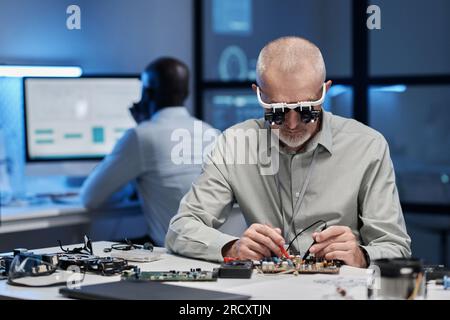 Erfahrener Ingenieur in Spezialgläsern, der sich auf seine Arbeit mit einem Computerchip am Tisch im Labor konzentriert Stockfoto