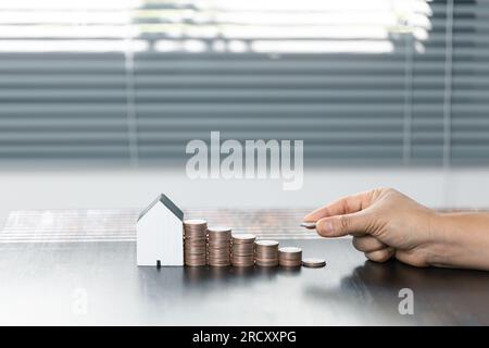 Geld sparen für Haus und Immobilien. Frau Hand Schutz auf Stapel Münzen und Hausmodell auf dem Tisch. Stockfoto