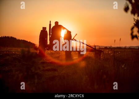 Bauer auf einem Traktor, kultiviert das Land, an einem heißen Tag, bei Sonnenuntergang. Silhouette vor dem Hintergrund der untergehenden Sonne Stockfoto