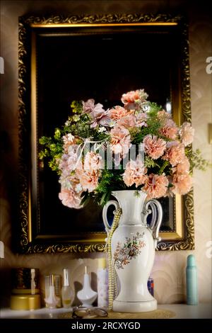 Künstliche Blumen in puderrosa Farbe stehen in einer hohen Vase vor einem Spiegel und auf einer Kommode. Rustikale Einrichtung im Retro-Stil. Stockfoto