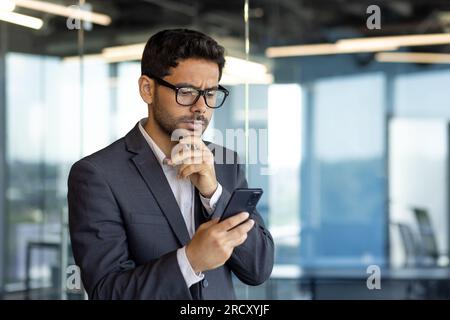 Ein Geschäftsmann, der im Büro telefoniert, arbeitet mit einer arabischen Brille am Arbeitsplatz und nutzt die App auf dem Smartphone, um Internetseiten durchdacht zu lesen und zu durchsuchen. Stockfoto