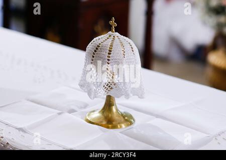 goldener MetallKelch oder Becher mit Symbol des Kreuzes Christi, bedeckt mit weißem, aufgesticktem Stoff auf dem Altar - Ampulle Stockfoto