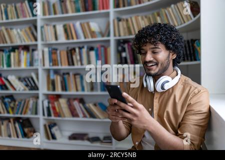 Ein junger hispanischer Schüler, der lächelt und glücklich ist, die Online-Lern-App nutzt, ein lächelnder Mann in Kopfhörern, der in der akademischen Bibliothek unter Büchern sitzt. Stockfoto