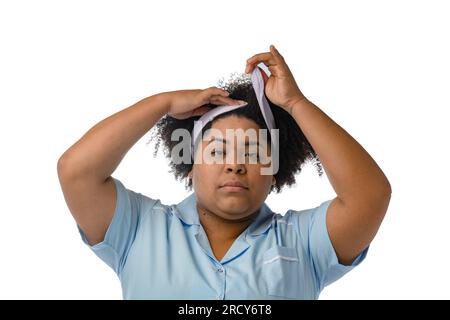 Junge Afro-Latina-brünette Frau, die ein Kopfband zur Gesichtsreinigung anlegt, in die Kamera schaut, weißer Hintergrund. Stockfoto