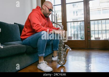 Ein junger lateinischer Musiker zu Hause, der auf dem Sofa sitzt, beruhigt nach unten schaut und denkt, sein Saxophon über dem Boden zu halten, Musikkonzept mit Kopierraum. Stockfoto