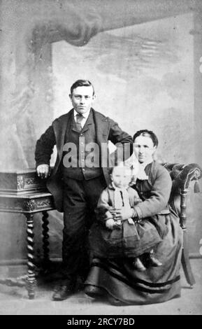 Porträt eines Mannes, der neben einer Dame steht, mit einem Kind auf dem Schoß. Originalfoto von Henry Howell, Kunststudio, Lammas Street, Carmarthen, Wales. Aus einer Sammlung viktorianischer Carte de Visite Fotografien aus den 1890er Jahren. Carte de visite ist ein französischer Begriff, der auf Englisch als „Visitenkarte“ bezeichnet wird. Normalerweise wurde ein Fotodruck aus Albumen von etwa 54 x 89 mm auf einer etwas größeren Karte angebracht. In einigen Fällen haben die Verwüstungen der Zeit ihren Tribut gefordert. Stockfoto