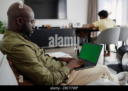 Ein afroamerikanischer Vater, der online an einem Laptop arbeitet, während er mit seinem Sohn auf dem Sofa sitzt und im Hintergrund Hausaufgaben macht Stockfoto