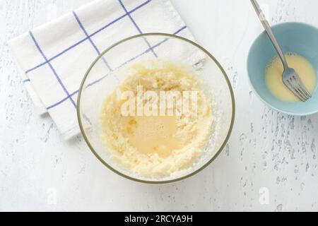 Glasschüssel mit Schlagsbutter mit Zucker und geschlagenem Ei auf hellblauem Hintergrund, Draufsicht. Hausgemachte Milchkuchen zubereiten, machen Sie es selbst, Schritt für Schritt Stockfoto