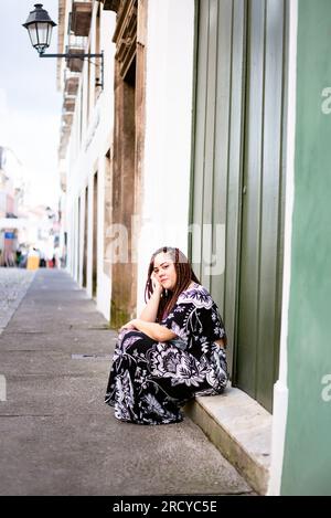 Eine schöne, entspannte Frau, die mit Zöpfen im Haar unterwegs ist und auf einer Stufe mit der Hand am Kinn sitzt. Alte Häuser im Hintergrund. Pelourinho, Salv Stockfoto