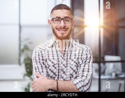 Junger, gutaussehender Geschäftsmann mit Brille, der mit gekreuzten Armen im Büro steht. Stockfoto