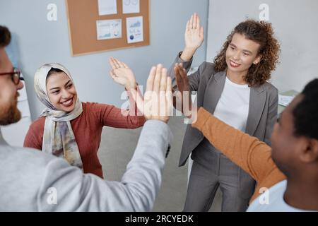 Fröhliche, junge interkulturelle Kollegen in eleganter Freizeitkleidung, die sich gegenseitig High Five geben, während sie im Büro im Kreis vor der Kamera stehen Stockfoto
