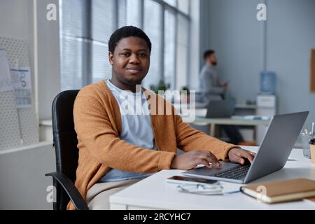Junger, selbstbewusster schwarzer Mann in Freizeitkleidung, der in die Kamera schaut, während er am Arbeitsplatz vor dem Laptop sitzt und Online-Informationen analysiert Stockfoto