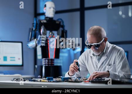 Erfahrener Ingenieur, der mit der Computerplatine eines Roboters arbeitet, während er an seinem Arbeitsplatz im Labor sitzt Stockfoto