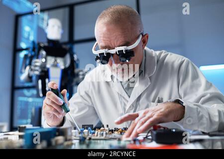 Ausgereifter Programmierer im Labormantel, der am Tisch sitzt und die Hauptplatine im Büro zusammensetzt Stockfoto