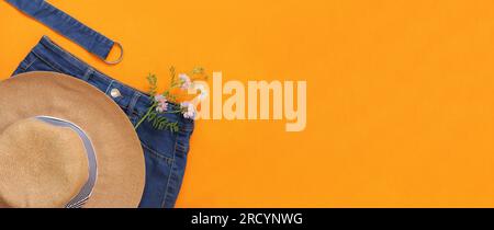 Sommerfoto mit Kleidung auf hellem orangefarbenen Hintergrund. Sommerhut, Jeansshorts und Jasminblumen. Speicherplatz kopieren. Draufsicht, flach liegend. Mode und Stylie Stockfoto