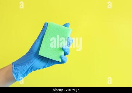 Die Hand einer Frau im Handschuh hält einen Schwamm zum Geschirrspülen. Hausreinigung. Hand mit einem grünen Schwamm auf gelbem Hintergrund. Reinigungsanzeige Stockfoto