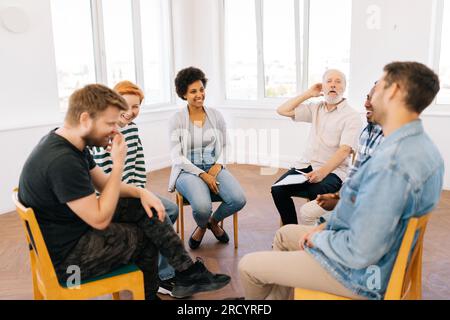 Weitwinkel einer fröhlichen interrassischen Gruppe von Menschen mit Alkoholabhängigkeit, die im Reha-Zentrum im Kreis während der Gruppentherapiesitzung sprechen. Stockfoto