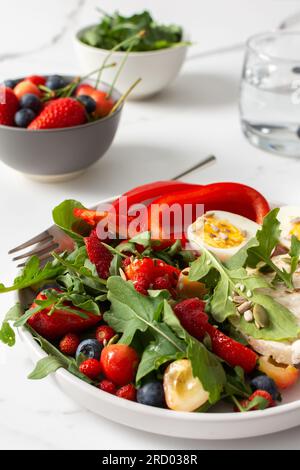 Gesundes, leckeres Frühstück, ein Teller mit gekochten Eiern und Reisbrot, Salat aus Rucola und Erdbeeren mit Kirschen und Blaubeeren, frischer Paprika Stockfoto