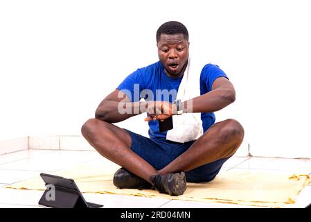 Porträt eines attraktiven jungen, sportlich überraschten Mannes, der auf dem Boden sitzt und die Zeit auf seiner Armbanduhr betrachtet. Stockfoto
