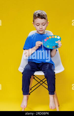 Kleiner netter Vorschuljunge, der eine Palette von Farben und einen Pinsel auf gelbem Hintergrund intensiv hält. Kreatives Kind. Stockfoto