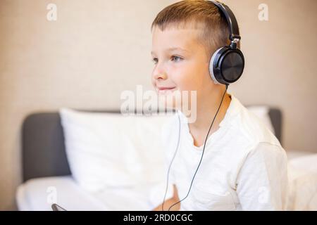 Lächelnder, glücklicher, weißer Junge im T-Shirt, der auf dem Bett sitzt und einen Tablet-Computer benutzt Stockfoto