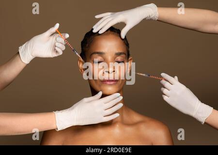 Attraktive afroamerikanische Frau mittleren Alters, die von zwei Kosmetikern Gesichtsspritzen bekommt, brauner Hintergrund Stockfoto