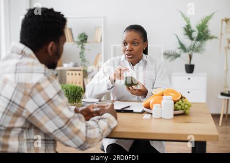 Konzentrieren Sie sich auf eine afroamerikanische Frau im Labormantel, die grüne Früchte hält, während sie am Arbeitsplatz des Arztes bleibt. Ernster Facharzt diskutiert Nutzen von Avocado in einer gesunden Ernährung mit aufmerksamen Mann. Stockfoto