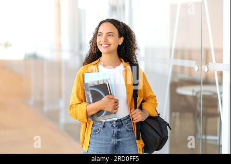 Weibliche Studentin. Glückliche, reizende brasilianische oder hispanische Studentin, mit einem Rucksack, mit Büchern und Notizbüchern in der Hand, steht in der Nähe des Universitätscampus, sieht und lächelt zur Seite Stockfoto