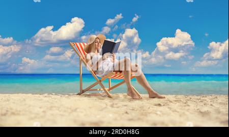 Junge Frau in einem weißen Kleid, die auf einem Liegestuhl sitzt und am Meer ein Buch liest Stockfoto