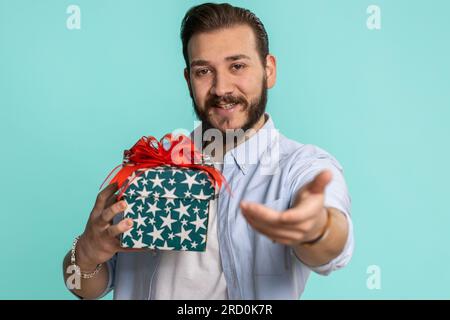 Ein positiv lächelnder junger libanesischer Mann, der eine Geburtstagsgeschenkbox überreicht, streckt sich die Hände aus, bietet einen eingepackten Geschenk Karriereprämie an, feiert Partyverkauf. Bärtiger arabier isoliert auf blauem Studiohintergrund Stockfoto