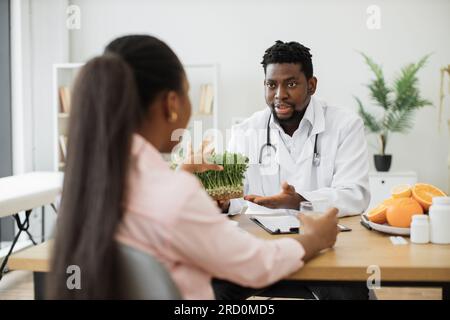 Schulterblick eines afroamerikanischen Mannes mit Stethoskop, der einer hübschen Frau auf der Arbeit Mikrokräuter vorschlägt. Facharzt für Lebensmittel mit jungen pflanzlichen Sprossen als gesundheitsfördernde Komponente. Stockfoto